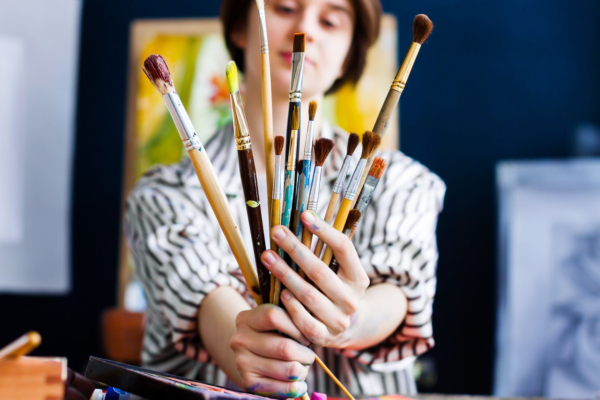 Young beautiful artist teacher student woman girl boy in art workshop studio surrounded by colorful black white painting hold huge bunch stack of art brushes for oil acrylic paints wear striped robe
